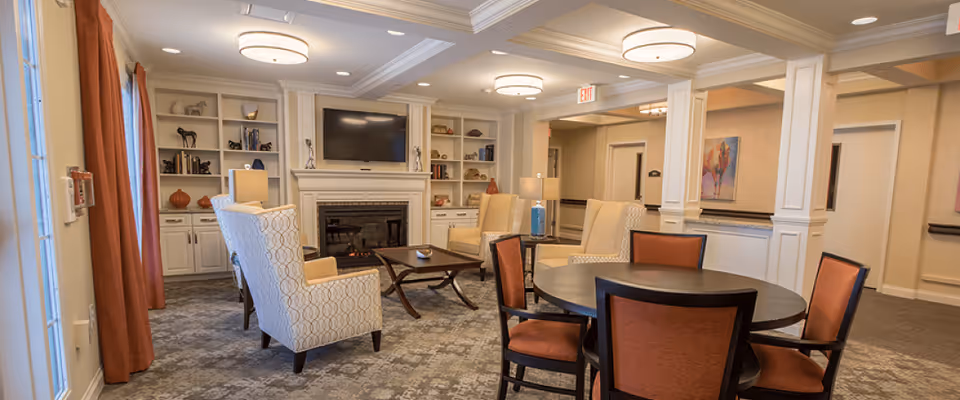 Well-lit common area with upholstered armchairs, a fireplace and TV, built-in shelving, and a round table with chairs.