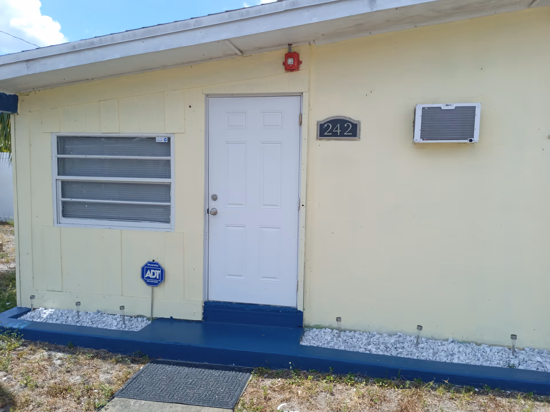 Exterior view of a single-story yellow building with a white door numbered 242, a window with horizontal blinds, a wall-mounted air conditioning unit, and a small garden bed with white rocks and solar lights in front.