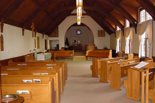 Interior view of a chapel with wooden pews arranged in rows facing an altar at the front. The ceiling is vaulted with wooden beams, and there are large windows with curtains on the right side allowing natural light to enter. The altar area has religious symbols including a cross and a stained glass window above.