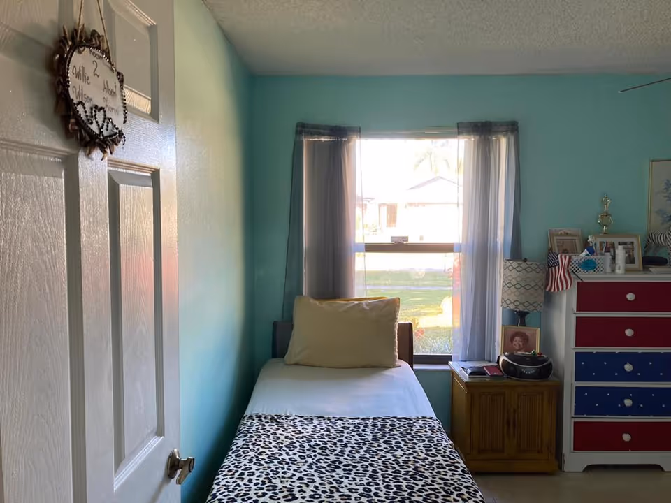 A small bedroom with light blue walls and a single bed covered with a white sheet and a leopard print blanket. There is a beige pillow on the bed. A window with sheer gray curtains lets in natural light. Next to the bed is a wooden nightstand with a lamp, a photo frame, and a radio. To the right, there is a dresser with red, white, and blue drawers, decorated with framed photos, a small American flag, and a trophy. The door to the room is partially visible with a heart-shaped hanging sign.