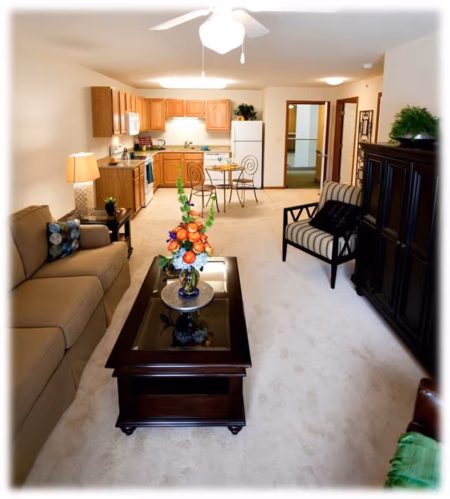 A bright and cozy living area in a retirement community apartment featuring a beige sofa with patterned pillows, a dark wooden coffee table with a floral arrangement, a striped armchair, and a dark wooden cabinet. The background shows a small kitchen with wooden cabinets, a white refrigerator, stove, and a round dining table with two chairs. The room is carpeted and well-lit with a ceiling fan light and a table lamp.