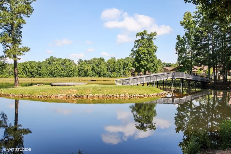 A serene outdoor scene featuring a small pond with clear reflections of trees and clouds, a wooden footbridge crossing over the water, and a grassy area with a stone border. Trees and a partly cloudy blue sky are visible in the background.