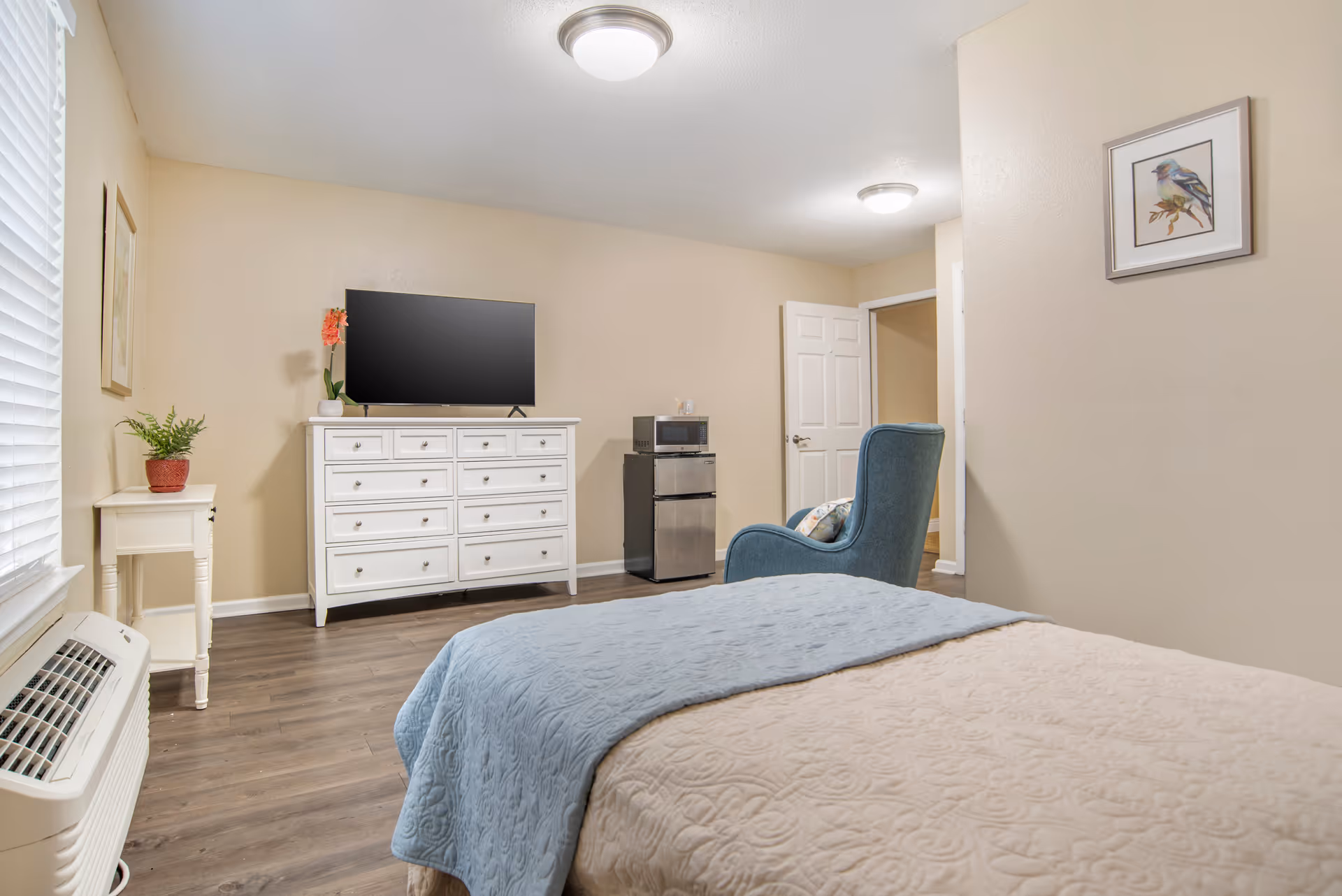 A cozy bedroom in a senior living facility featuring a bed with a beige quilt and blue blanket, a white dresser with a flat-screen TV on top, a small refrigerator and microwave, a blue upholstered chair, a small white side table with a plant, and framed artwork on the walls. The room has wood flooring and neutral-colored walls.