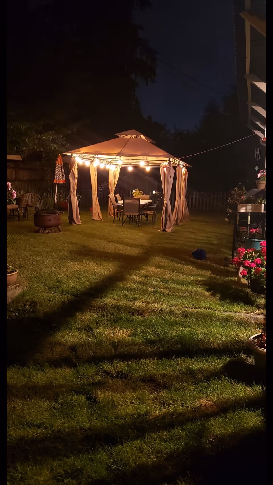 A nighttime outdoor scene showing a lit gazebo with string lights hanging around its roof. Inside the gazebo, there is a table with chairs. The surrounding yard has green grass, potted plants with flowers, and a fire pit to the left. Trees and a fence are visible in the background.