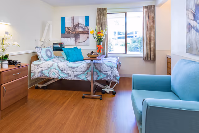 A bright and clean rehabilitation center room featuring a hospital bed with patterned bedding and teal pillows, a small overbed table with a vase of flowers, a wooden cabinet with a phone and orchid plant, a teal armchair, and a large window with curtains letting in natural light.