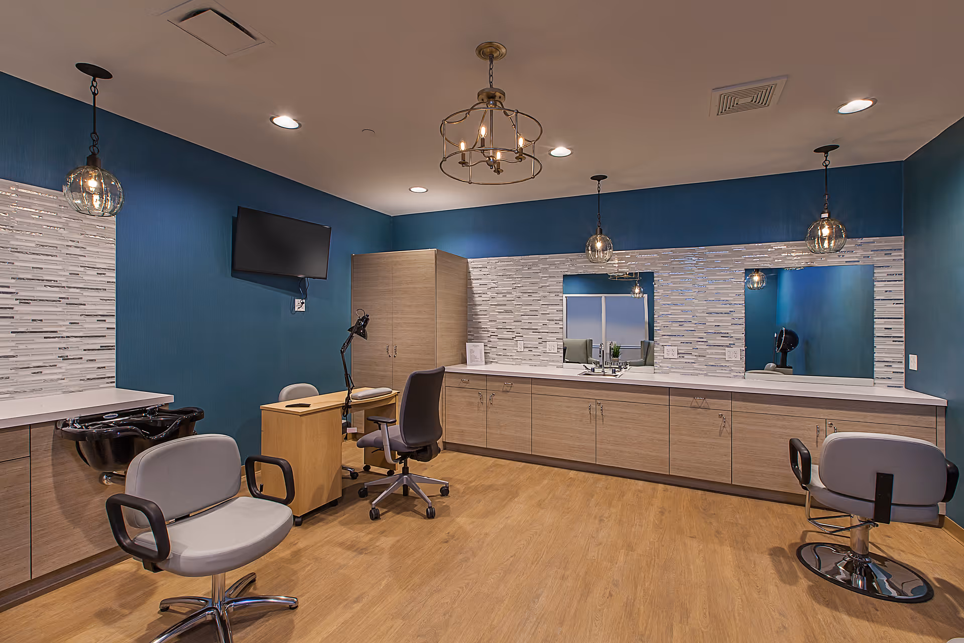 Interior view of a modern salon area with blue walls, wooden flooring, and a long counter with cabinets and two large mirrors. There are two salon chairs, a small desk with an office chair, a wall-mounted TV, and three pendant lights hanging from the ceiling.