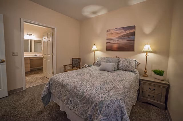 A cozy bedroom with a double bed covered in a patterned comforter and pillows. There are two bedside tables with lamps on either side of the bed, a wooden chair with a cushion, and a framed picture of a beach scene hanging on the wall above the bed. An open door reveals a bathroom with a sink and mirror.