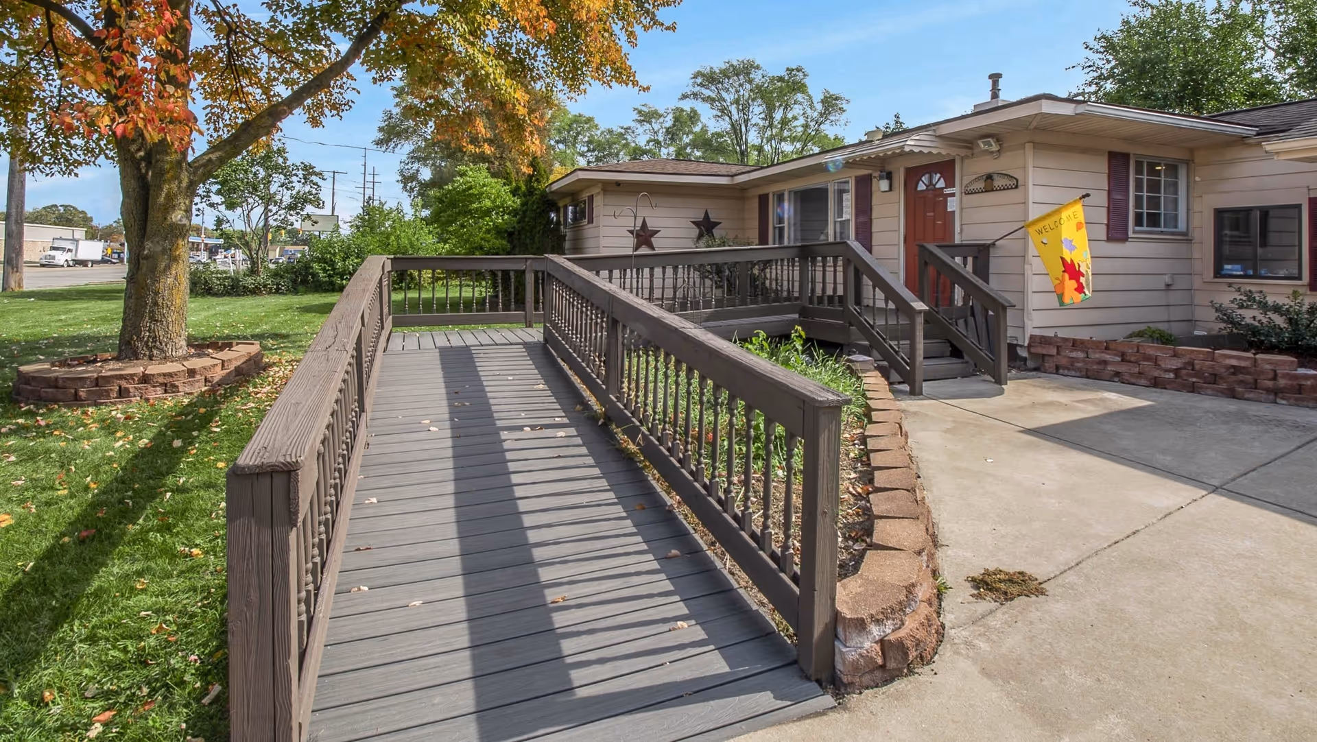 Exterior front entrance of a single-story assisted living building with a wooden wheelchair ramp leading to the front door and a 'Welcome' flag.