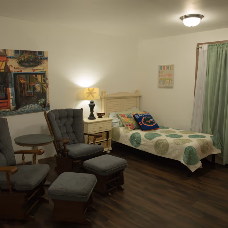 A cozy bedroom with a single bed covered with a white quilt featuring green circular patterns. The bed has colorful pillows, including one with a Florida Gators logo. Next to the bed is a small white nightstand with a lamp and a small decorative bowl. Two cushioned wooden rocking chairs with matching footstools are positioned near a small round table. The room has wooden flooring, a window with green curtains, and wall art including a painting of a street scene and a framed inspirational quote.