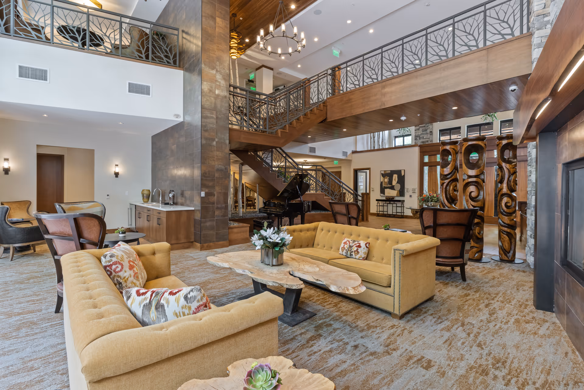 Spacious and elegantly decorated living room area in a senior living facility featuring two mustard yellow sofas with patterned cushions, a natural wood coffee table with a floral centerpiece, a grand piano near a staircase with decorative metal railings, and wooden carved pillars. The room has a high ceiling with modern chandeliers, a fireplace on the right, and a carpeted floor with earth tones.