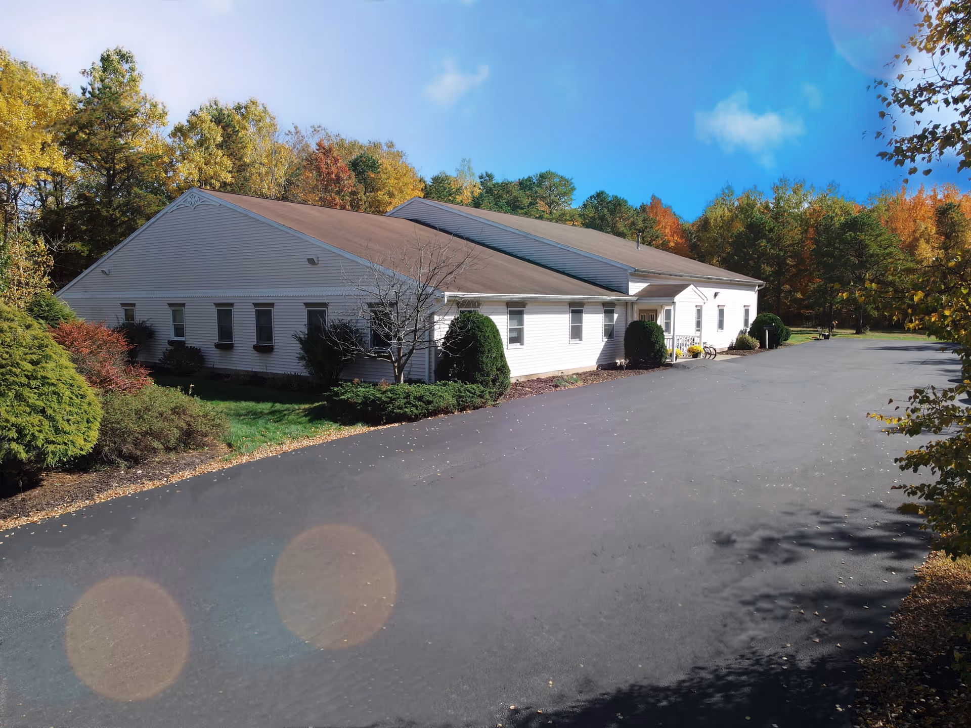 Exterior view of a single-story white building with a brown roof surrounded by trees with autumn foliage under a clear blue sky. The building is set next to a paved driveway or parking area with some landscaping including bushes and small trees.