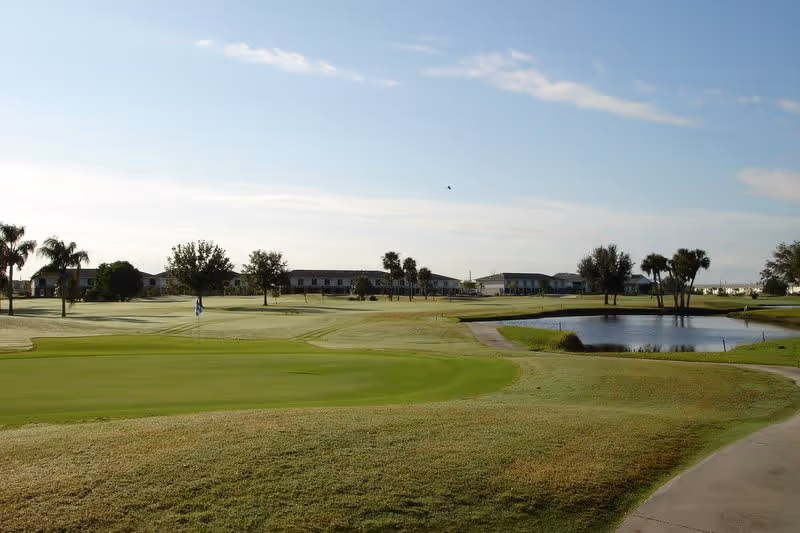 A scenic view of a golf course with well-maintained green grass, a putting green with a flag, a small pond, and several palm trees. In the background, there are low-rise buildings under a partly cloudy sky.