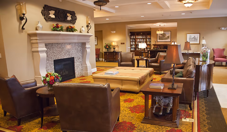 A cozy living room area in a retirement community featuring a large stone fireplace, leather armchairs, a leather sofa, a large ottoman, side tables with lamps, floral arrangements, and a colorful patterned carpet. In the background, there is a bookshelf filled with books and framed artwork on the walls.