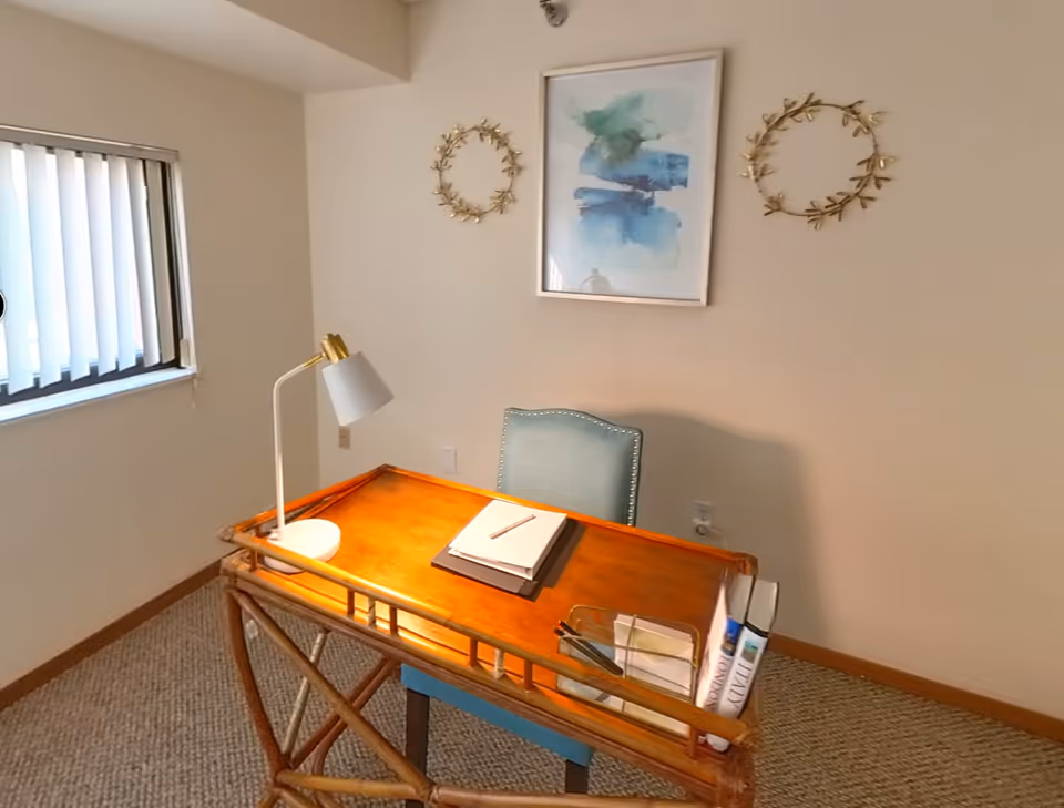 A small office or study area with a wooden desk and a blue upholstered chair. On the desk are a white desk lamp, a closed notebook with a pencil on top, and a few books held in a clear organizer. The wall behind the desk has two decorative gold wreaths and a framed abstract watercolor painting in shades of blue and green. A window with vertical blinds is on the left side of the room.