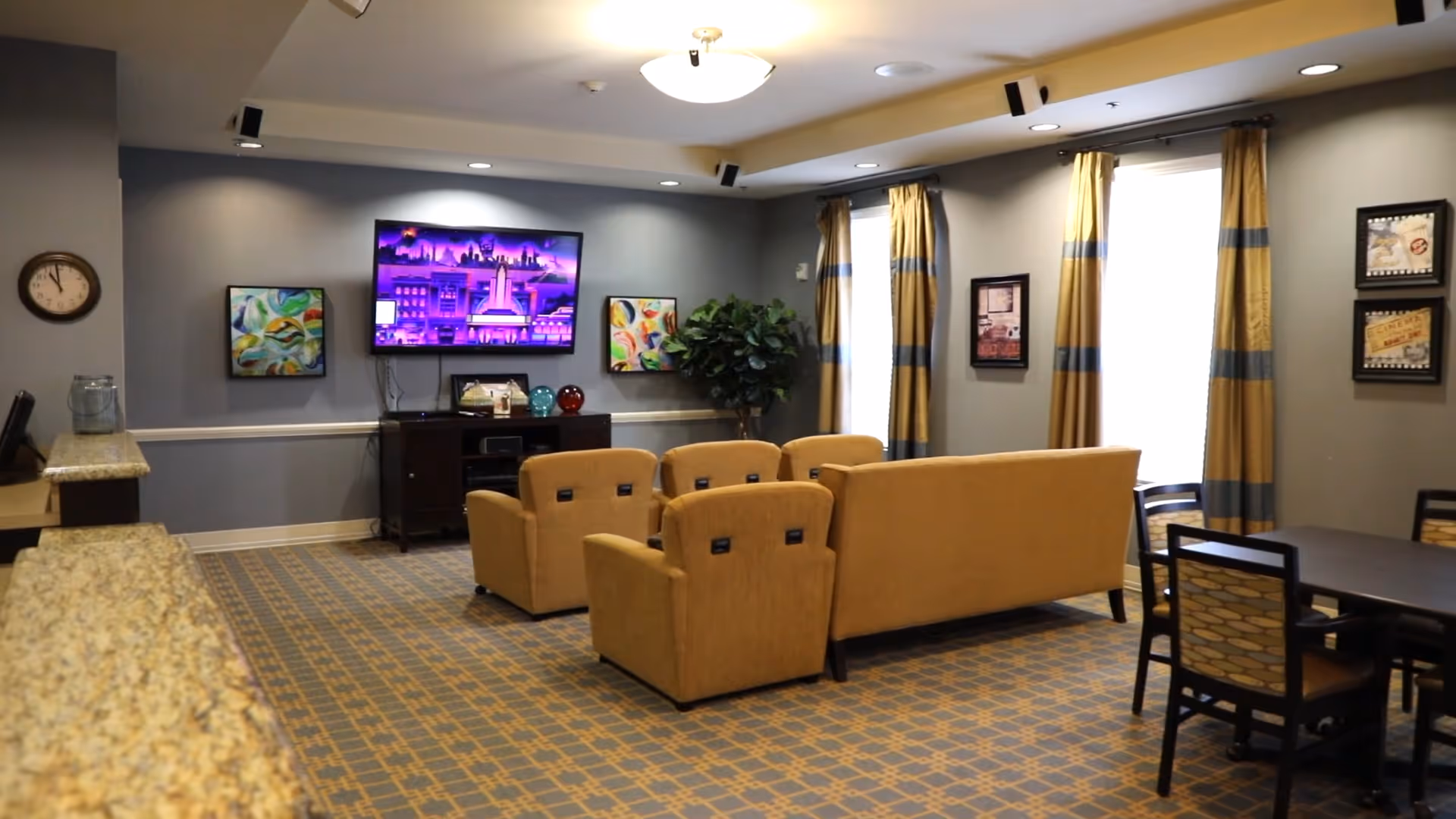 Comfortable senior living common room with a wall-mounted TV, upholstered chairs and sofa, a dining table, and patterned carpet.