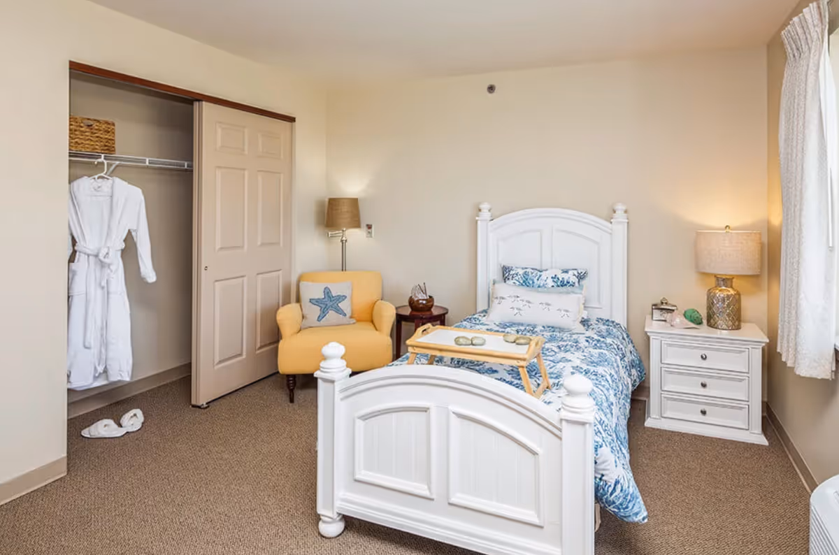 A bright bedroom with a white single bed dressed in blue floral bedding, a nightstand and lamp, a yellow armchair, and an open closet with a robe.