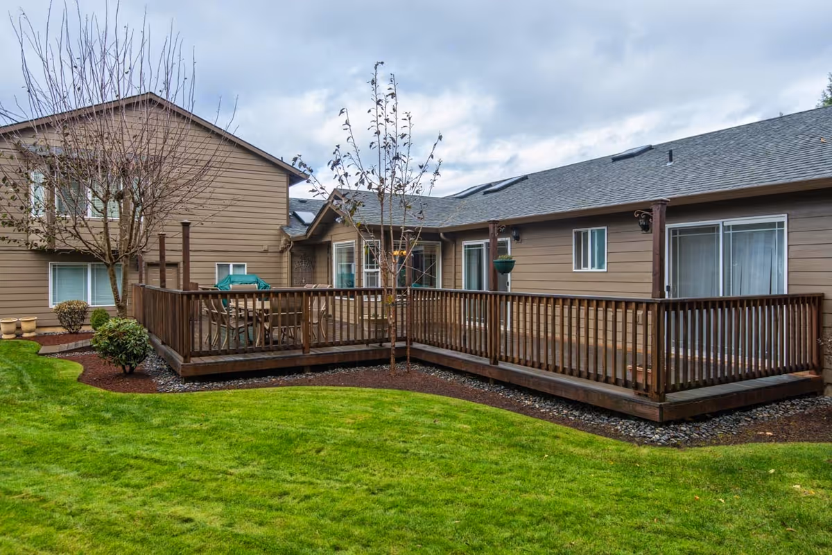 Exterior of a brown single-story adult care home featuring a wooden deck with outdoor furniture and a well-kept green lawn.
