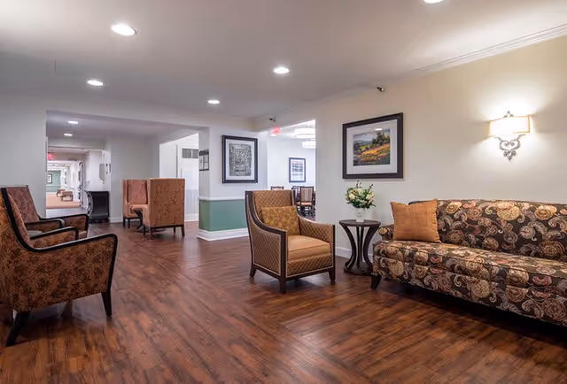 A spacious and well-lit common area with wooden flooring, featuring a patterned sofa with a brown cushion, two upholstered armchairs, a small round table with a flower arrangement, framed artwork on the walls, and a wall-mounted light fixture.