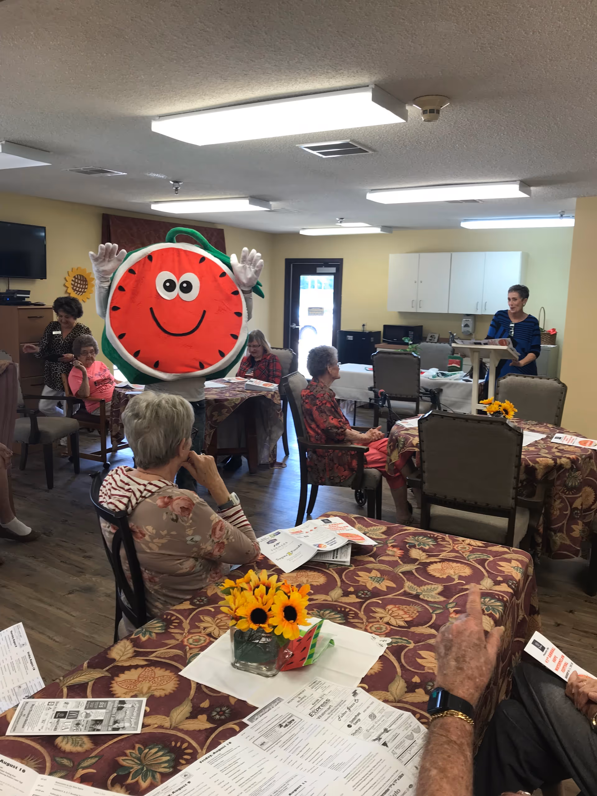 Residents seated at decorated tables in a communal dining/activity room as someone in a smiling watermelon mascot costume waves while a staff member speaks.