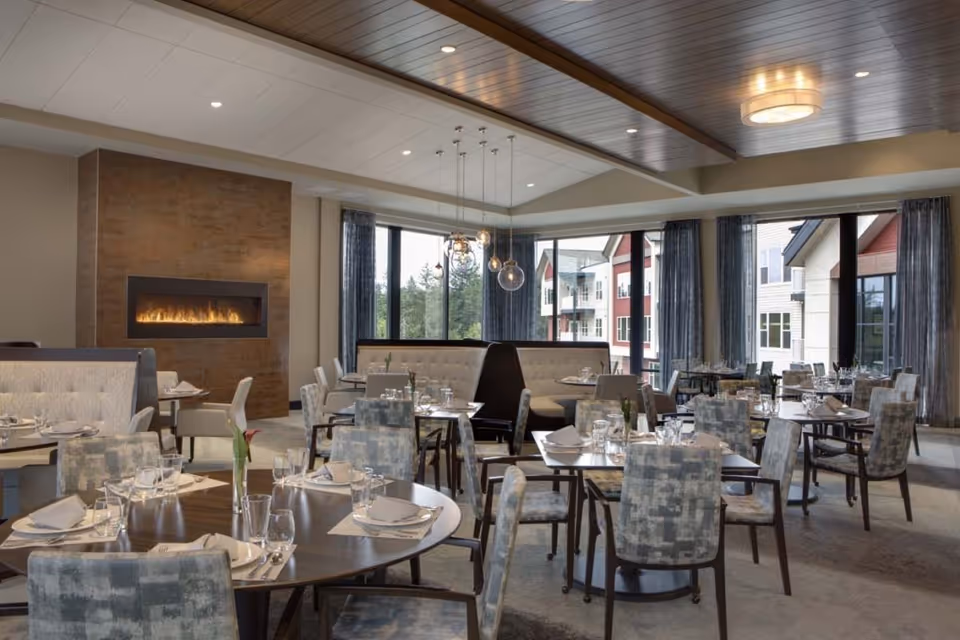 A spacious dining room with multiple round and rectangular tables set with plates, glasses, and napkins. The room features upholstered chairs and booth seating, large windows with curtains letting in natural light, and a modern fireplace on one wall. The ceiling has recessed lighting and a wooden panel design.