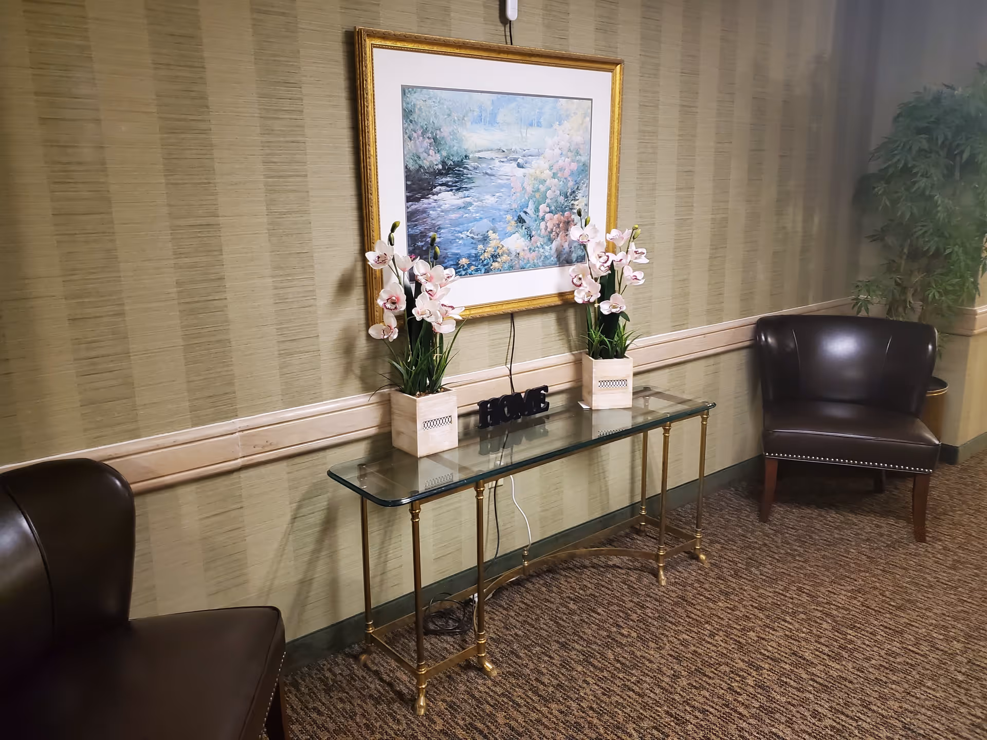 A cozy interior corner with two dark brown leather chairs facing each other, a glass-top table with gold legs between them, decorated with two potted orchid plants and a small decorative 'HOME' sign. Above the table hangs a framed painting of a serene river landscape. The walls have a striped beige wallpaper and the floor is carpeted in a brown pattern. A tall green plant is visible in the corner.