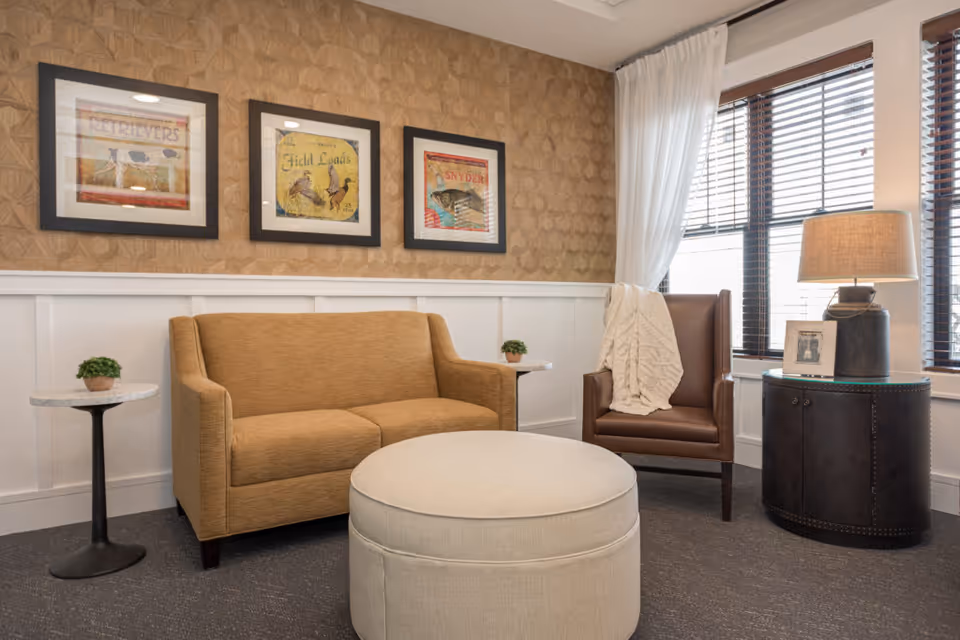 A cozy living room area with a tan sofa, a brown leather armchair draped with a white knitted blanket, a round beige ottoman, and two small side tables with potted plants. The wall behind the sofa features three framed vintage-style bird prints. A window with wooden blinds and white curtains allows natural light into the room, and a table lamp sits on a dark round cabinet beside the armchair.