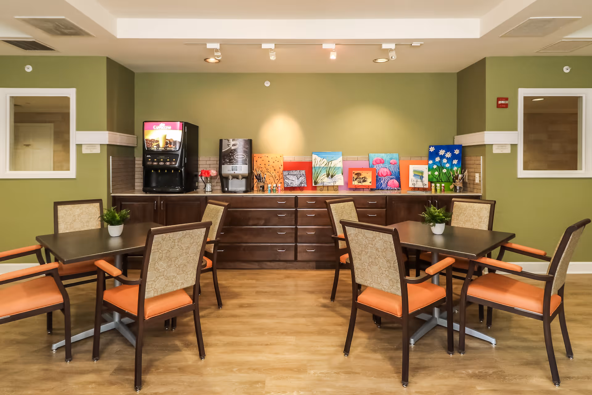 A dining area with two square tables, each surrounded by four chairs with orange seats and patterned backs. On each table is a small potted plant. Against the green wall at the back is a long dark wooden cabinet with multiple drawers and two beverage machines on top. Several colorful paintings are displayed on the cabinet. The floor is wooden, and there are two windows on the green walls on either side.