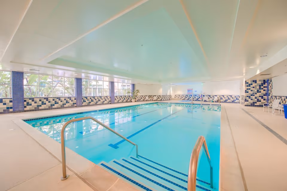 Indoor swimming pool with clear blue water, tiled walls in blue and white patterns, metal handrails leading into the pool, large windows letting in natural light, and a clean, spacious pool deck area with chairs.