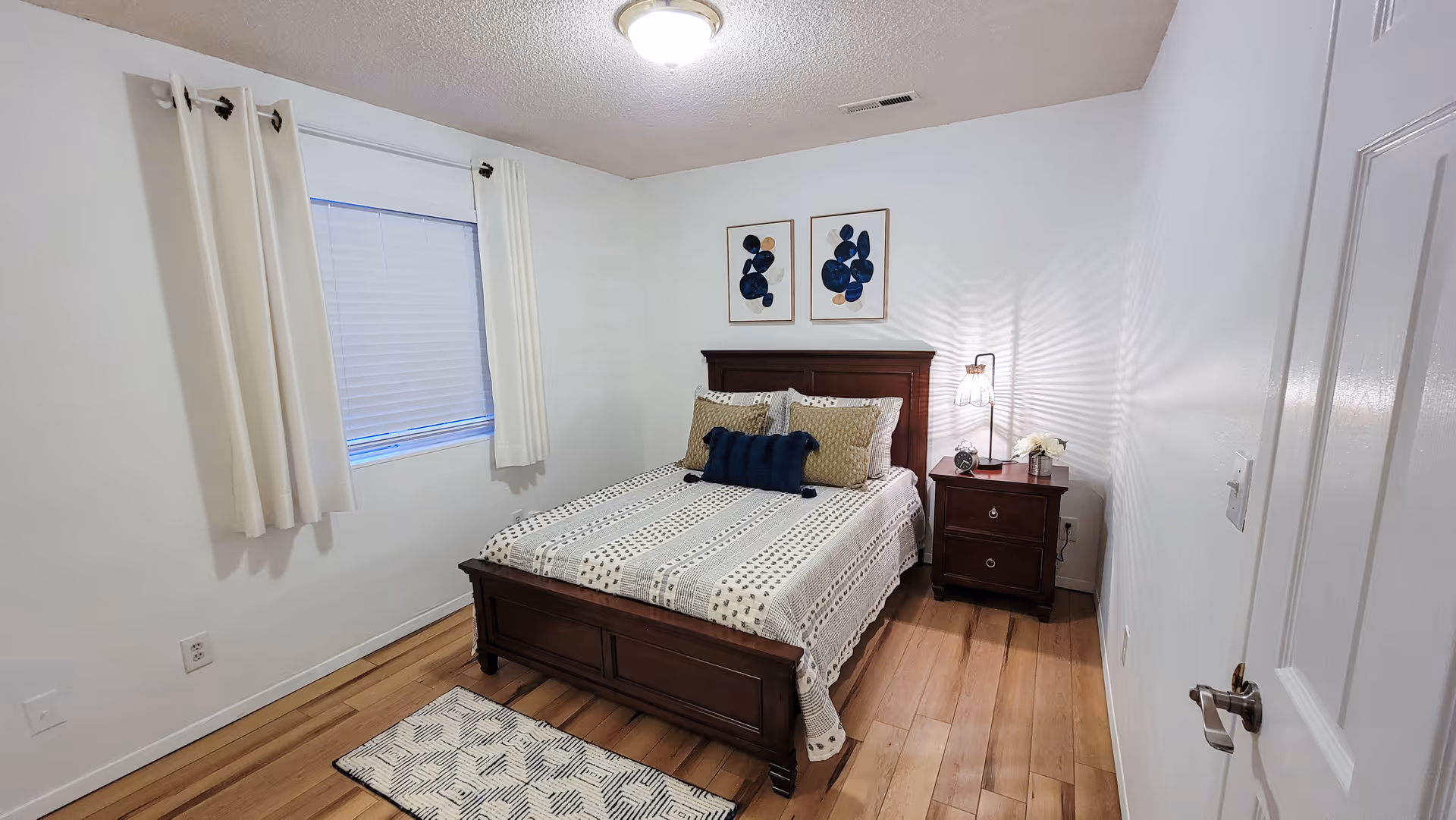 A small, neatly arranged bedroom with a wooden bed frame and a bed made with a patterned bedspread and multiple pillows. There is a wooden nightstand with a lamp, a clock, and a small flower vase. Two abstract art pieces hang on the wall above the bed. The room has light-colored walls, a window with white curtains, and wooden flooring with a small rug near the bed.