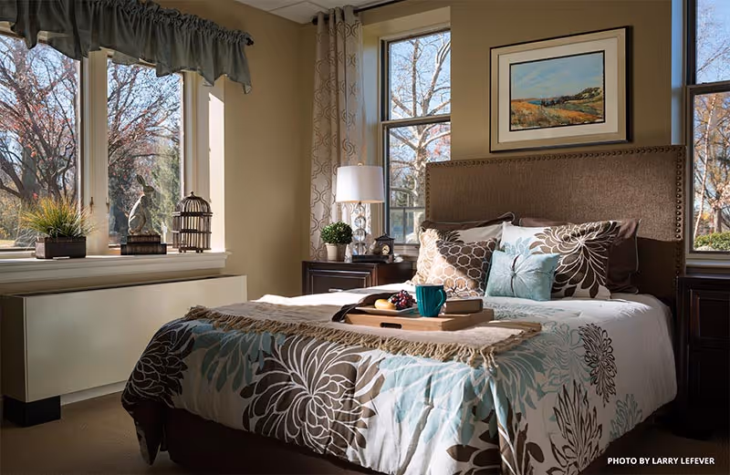 A cozy bedroom with a large bed featuring floral patterned bedding in shades of brown, blue, and white. The bed has multiple pillows and a tray with a cup and fruit on it. There are two windows with beige curtains letting in natural light, a nightstand with a lamp and a small plant, and a framed landscape painting on the wall above the headboard.