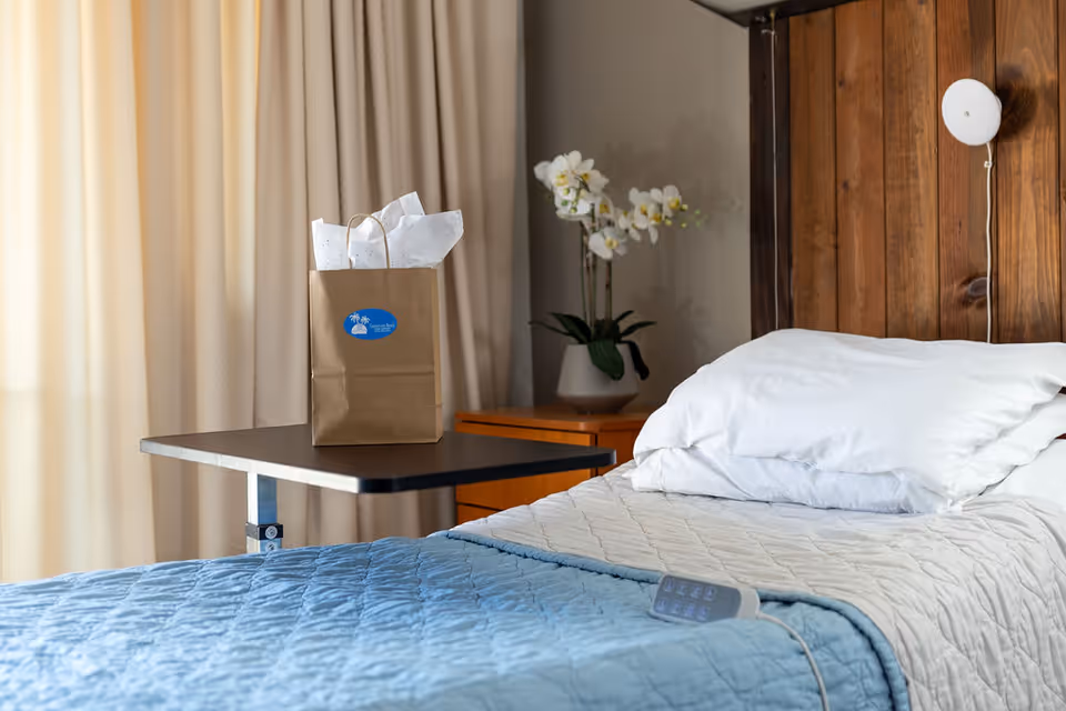 A neatly made bed with white pillows and a blue quilted blanket in a room with wooden paneling on the wall. A brown paper gift bag with white tissue paper sits on a black overbed table next to the bed. A small wooden nightstand with a white orchid plant in a pot is visible in the background, along with beige curtains.