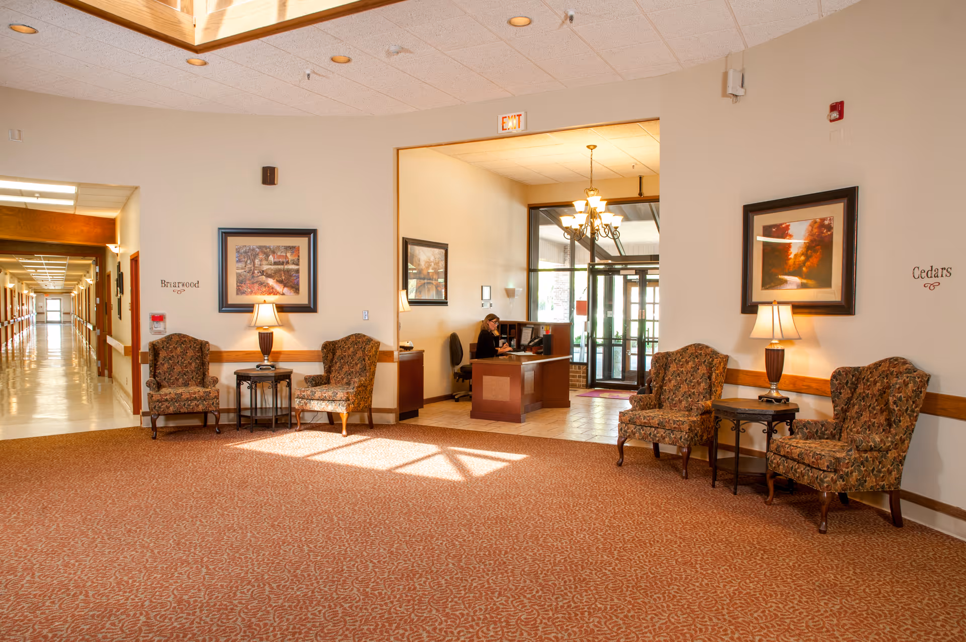 A spacious and well-lit senior living facility lobby area with patterned carpet flooring, four floral upholstered armchairs arranged in pairs on either side of two small tables with lamps. There are framed landscape paintings on the walls above the chairs. To the right, a receptionist is seated at a desk in a small office area with large windows and a glass door entrance. The hallway to the left leads to other rooms, with signs on the walls indicating 'Briarwood' and 'Cedars' sections.