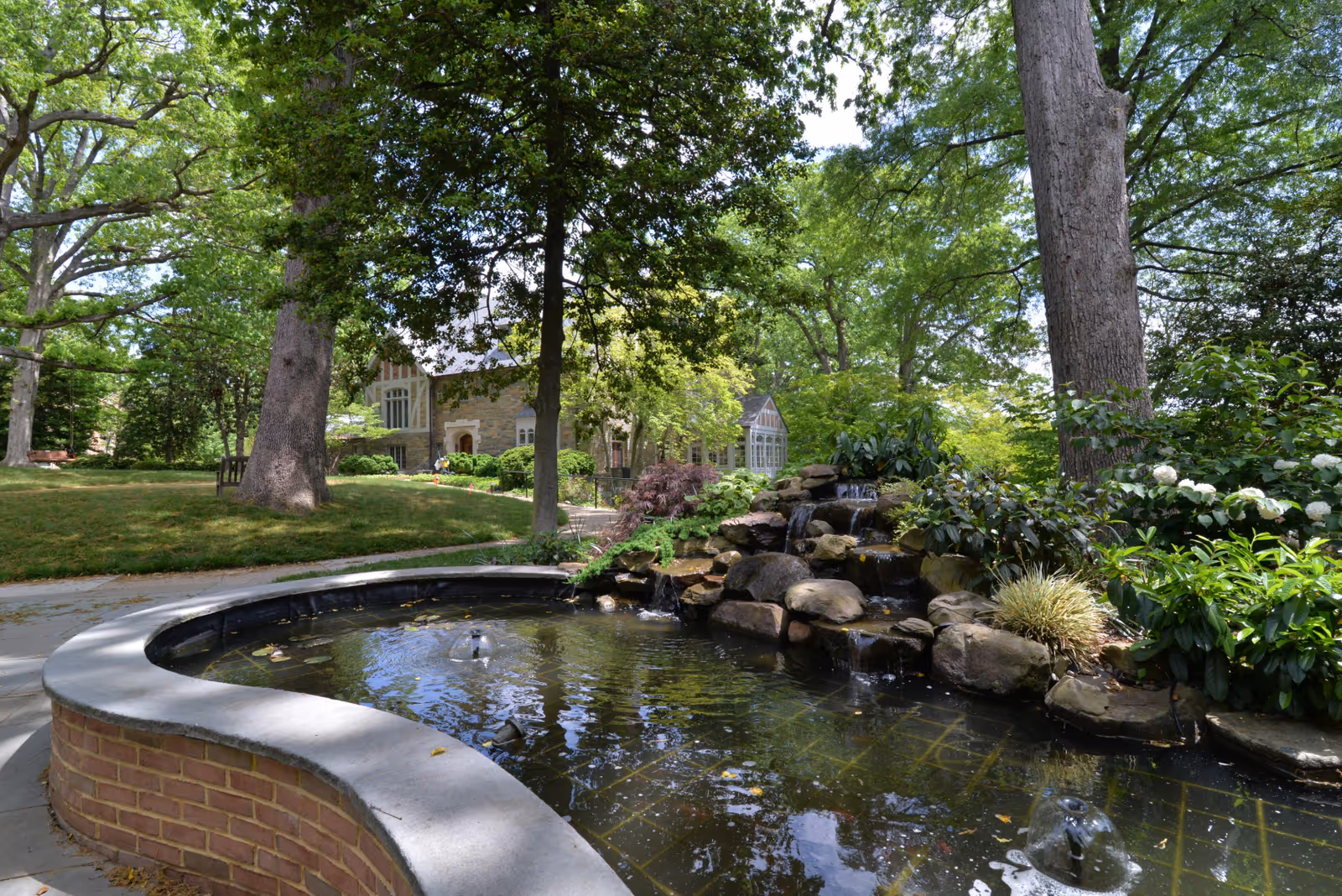 A serene outdoor garden area at Knollwood Military Retirement Community featuring a curved brick and stone water fountain with a small waterfall surrounded by lush green trees, shrubs, and flowering plants. A large building with stone walls and windows is visible in the background.