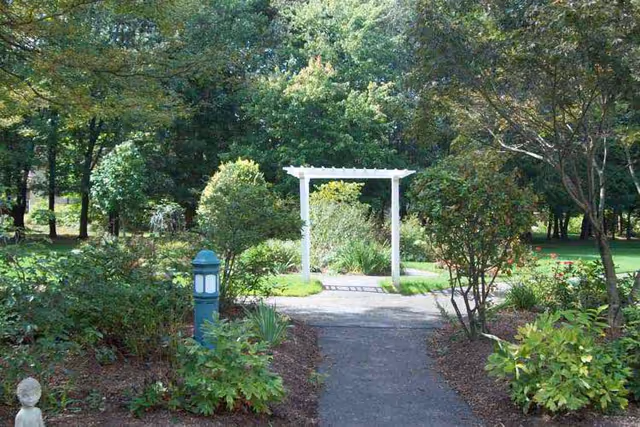 A garden pathway leading to a white wooden arbor surrounded by green bushes, trees, and various plants under a sunny sky.