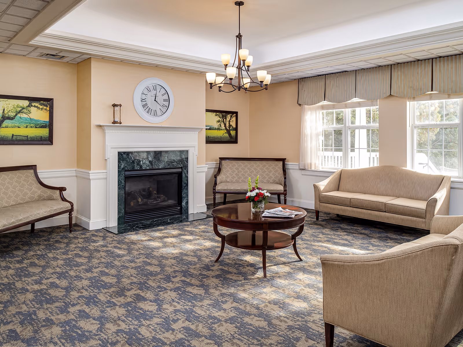 Bright communal living room with sofas, armchairs, a central coffee table, and a fireplace beneath a wall clock.
