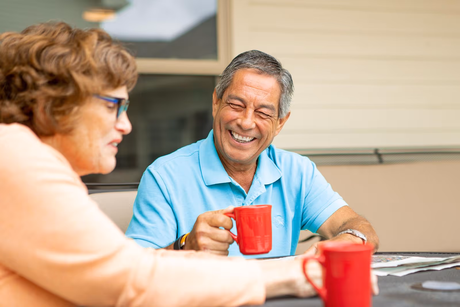 An elderly man and woman sitting outdoors at a table, smiling and holding red mugs, enjoying a conversation. The background shows a window and beige siding of a building.