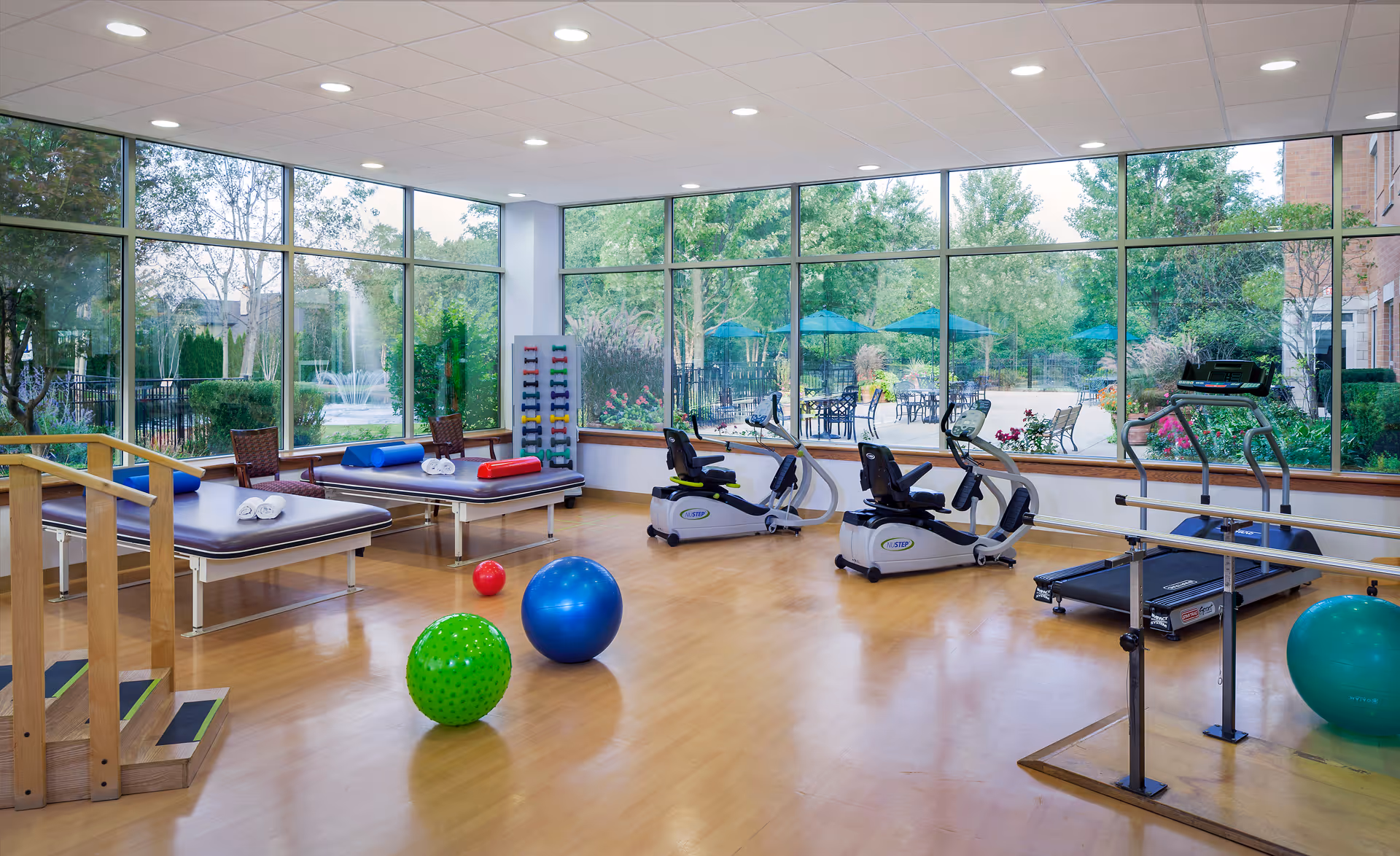 A bright and spacious exercise room with large floor-to-ceiling windows showing a garden outside. The room contains exercise equipment including two recumbent bikes, a treadmill, therapy tables with rolled towels, colorful exercise balls, a rack of small dumbbells, and wooden stairs with handrails for physical therapy.