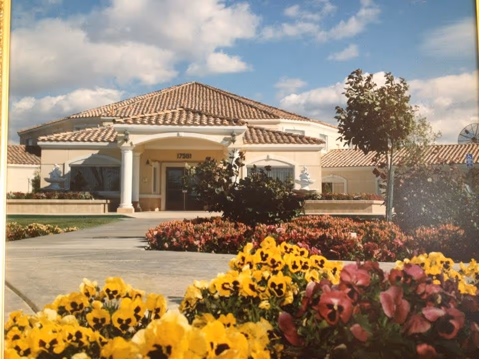 Front exterior view of Foremost Senior Campus building with a tiled roof, white columns at the entrance, and a well-maintained garden with colorful flowers and a tree in the foreground under a partly cloudy sky.