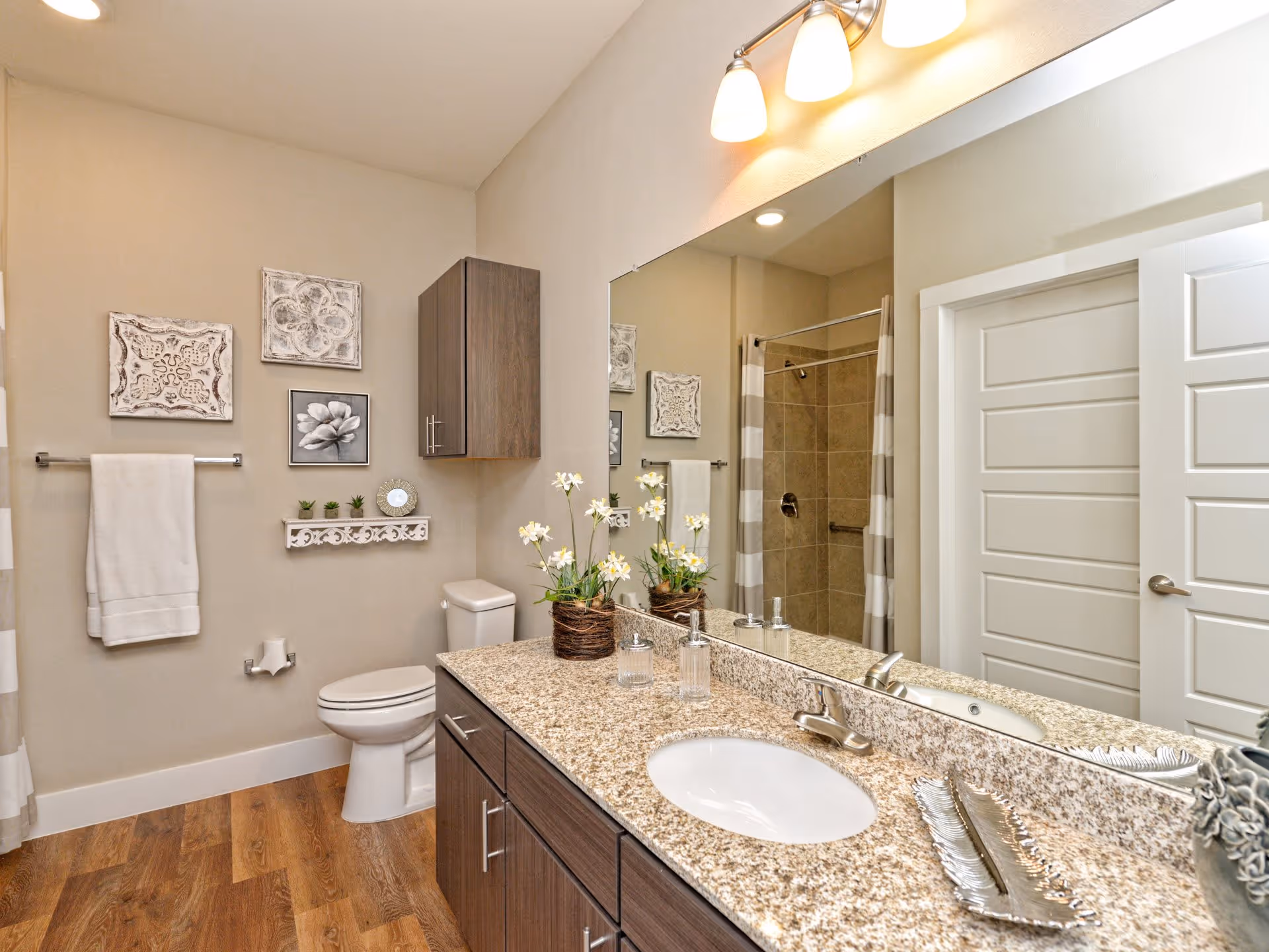 A modern bathroom with a granite countertop featuring a sink, soap dispenser, and decorative items. There is a large mirror above the countertop with three light fixtures. The bathroom has a toilet, a towel rack with a white towel, wall art, a small wall-mounted cabinet, and a shower with a striped curtain. The floor is wooden.