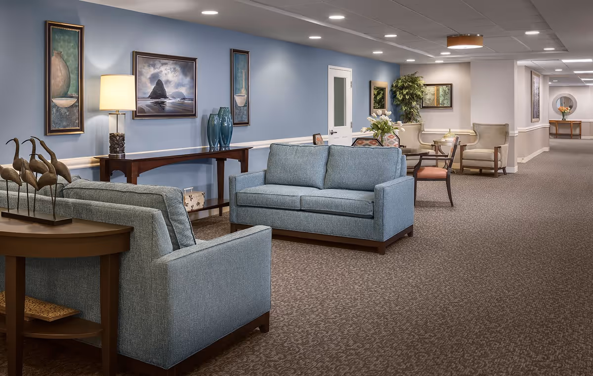 A spacious and well-lit senior living facility common area with blue upholstered sofas, armchairs, a wooden console table with decorative vases, framed artwork on the walls, and a carpeted floor. The hallway extends into the distance with additional seating and decorative plants.