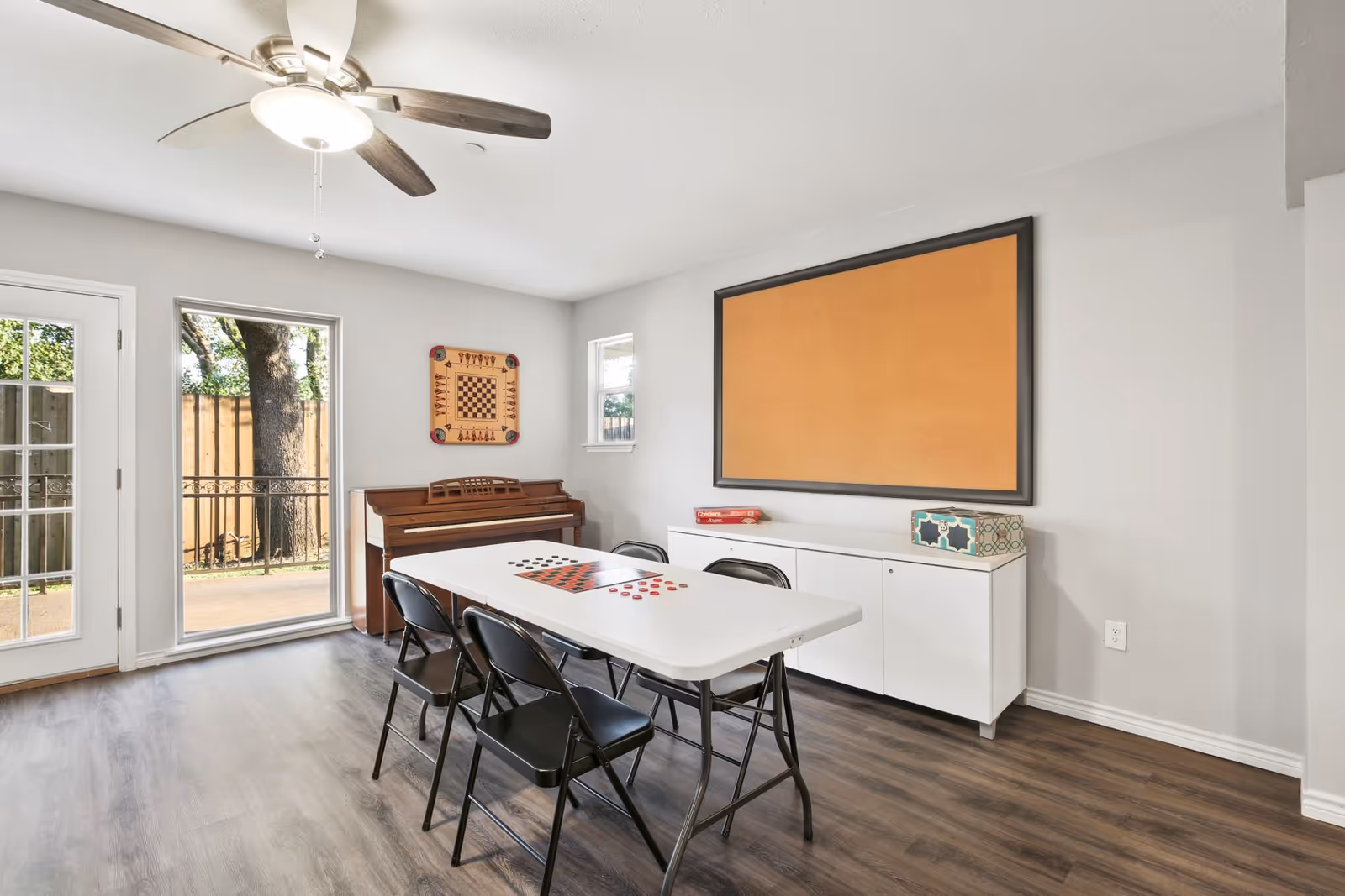 A bright room with a white folding table and four black folding chairs arranged around it. On the table are game boards for checkers and chess. Against the wall is a white cabinet with a large empty cork bulletin board above it. A piano is positioned in the corner beneath a wall-mounted chessboard. The room has wood flooring, a ceiling fan with light, and a glass door and window showing an outdoor area with trees and a wooden fence.