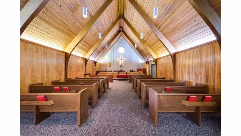 Interior view of a chapel with wooden pews arranged in two rows facing an altar with a cross on the wall. The ceiling is vaulted with wooden paneling and pendant lights hanging down. The walls are also wood-paneled and there is carpeted flooring.