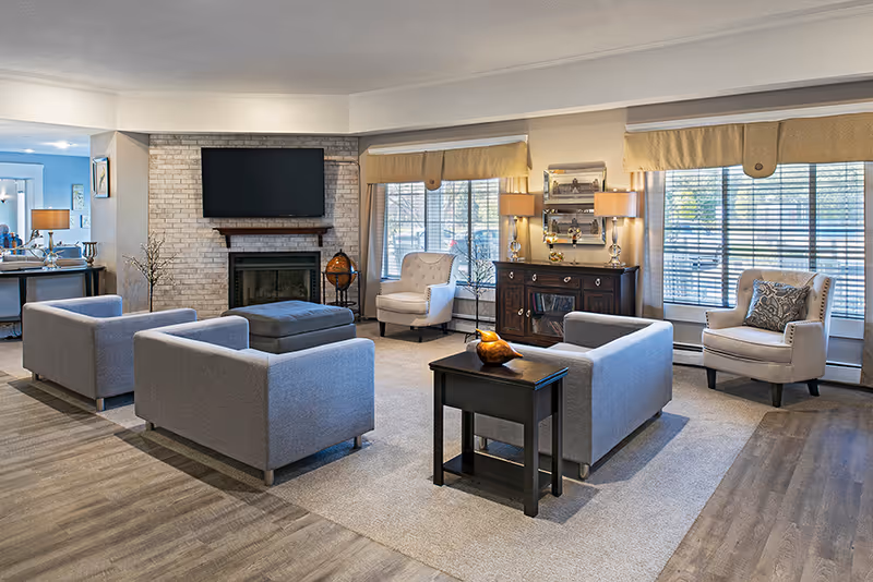 A spacious and well-lit senior living facility common area featuring a seating arrangement with two gray sofas, two beige armchairs, a dark wooden side table, and a large ottoman in the center. The room has large windows with blinds and beige valances, a brick fireplace with a mounted flat-screen TV above it, and a dark wooden cabinet with two lamps and decorative items. The floor is a combination of wood and carpet.