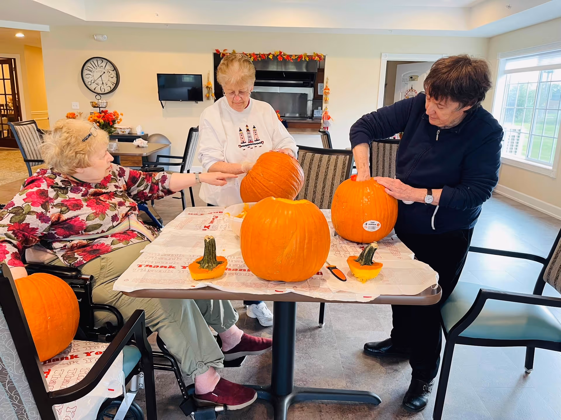 Three elderly women carving pumpkins at a table covered with newspaper in a well-lit room with large windows. One woman is seated in a wheelchair, while the other two are standing. The room has a clock on the wall, a TV, and fall-themed decorations.