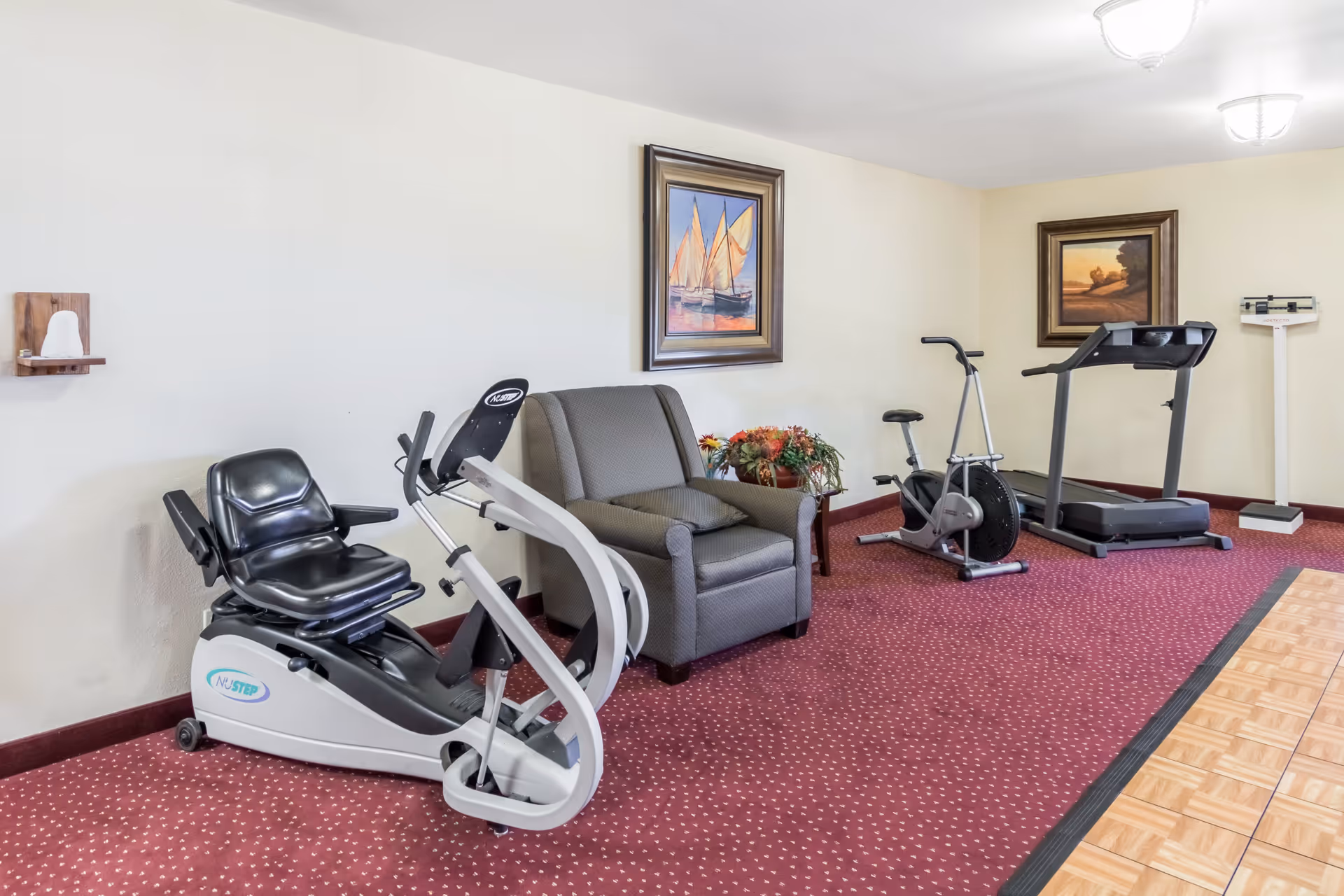 A small exercise room with a recumbent exercise bike, an upright exercise bike, and a treadmill. There is a gray armchair between the recumbent bike and the upright bike, with a small table holding a flower arrangement next to it. The room has red patterned carpet and light-colored walls with two framed paintings hanging on them. A weighing scale is visible in the back corner.