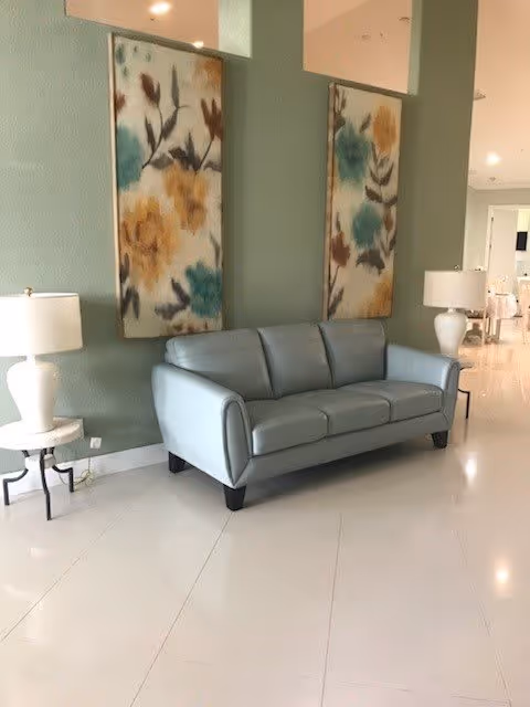 A modern interior space featuring a light blue leather sofa against a green wall with two large floral paintings. There are two white table lamps on either side of the sofa placed on small round tables. The floor is white and glossy, and the background shows a glimpse of another room with more furniture.