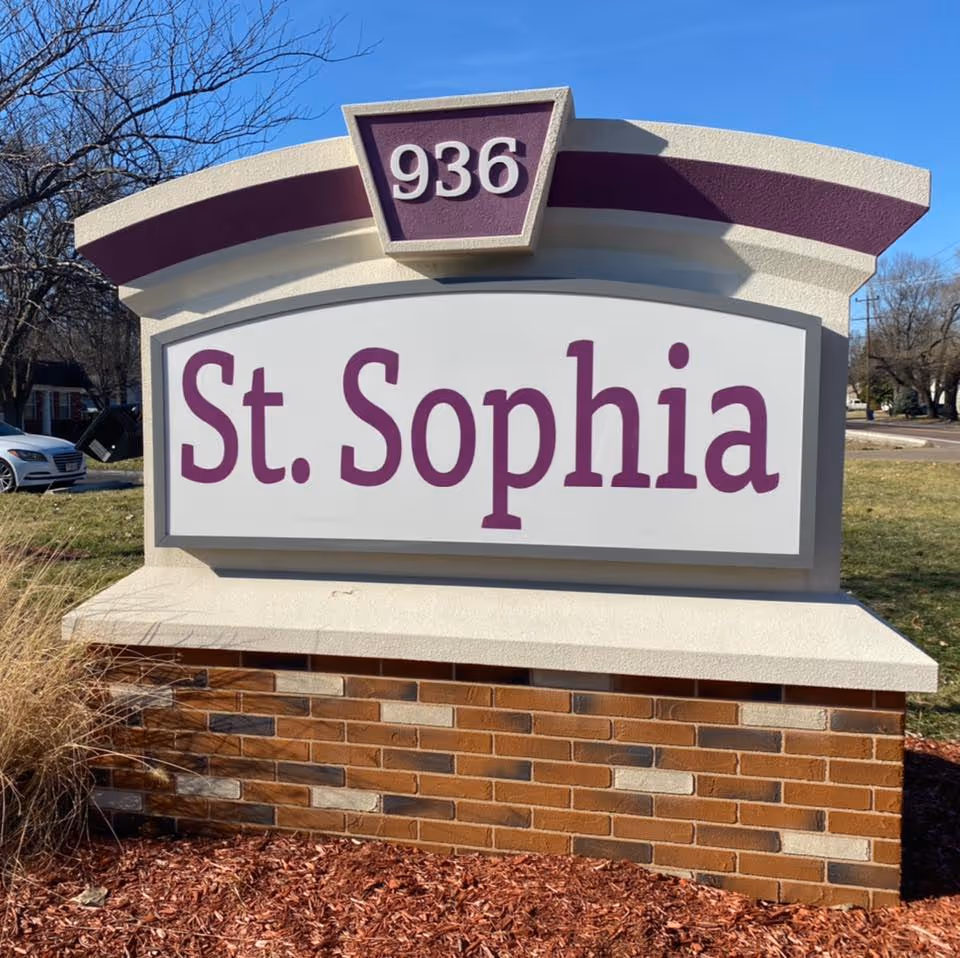 Outdoor sign for St. Sophia Health & Rehabilitation Center with the number 936 displayed above the name. The sign is mounted on a brick base with landscaping around it and a clear blue sky in the background.