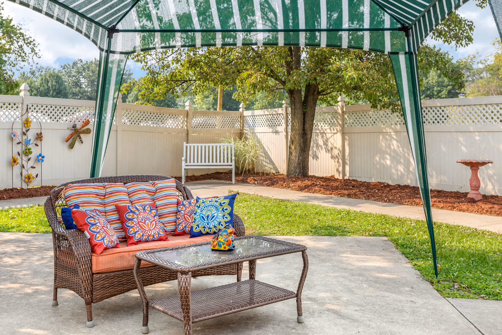 Outdoor patio area with a wicker loveseat adorned with colorful patterned cushions and a glass-top wicker coffee table under a green and white striped canopy. The area is surrounded by a white fence, with a tree, a white bench, decorative wall flowers, and a birdbath visible in the background.