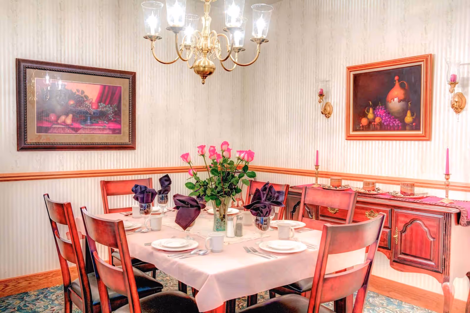 A formal dining room with a rectangular table covered with a white tablecloth. The table is set with white plates, cups, silverware, and dark purple folded napkins. A vase with pink roses is placed in the center of the table. The room has wooden chairs with dark cushions, a wooden sideboard with decorative candles and glass holders, and two framed paintings of fruit on the walls. A brass chandelier with glass shades hangs above the table.