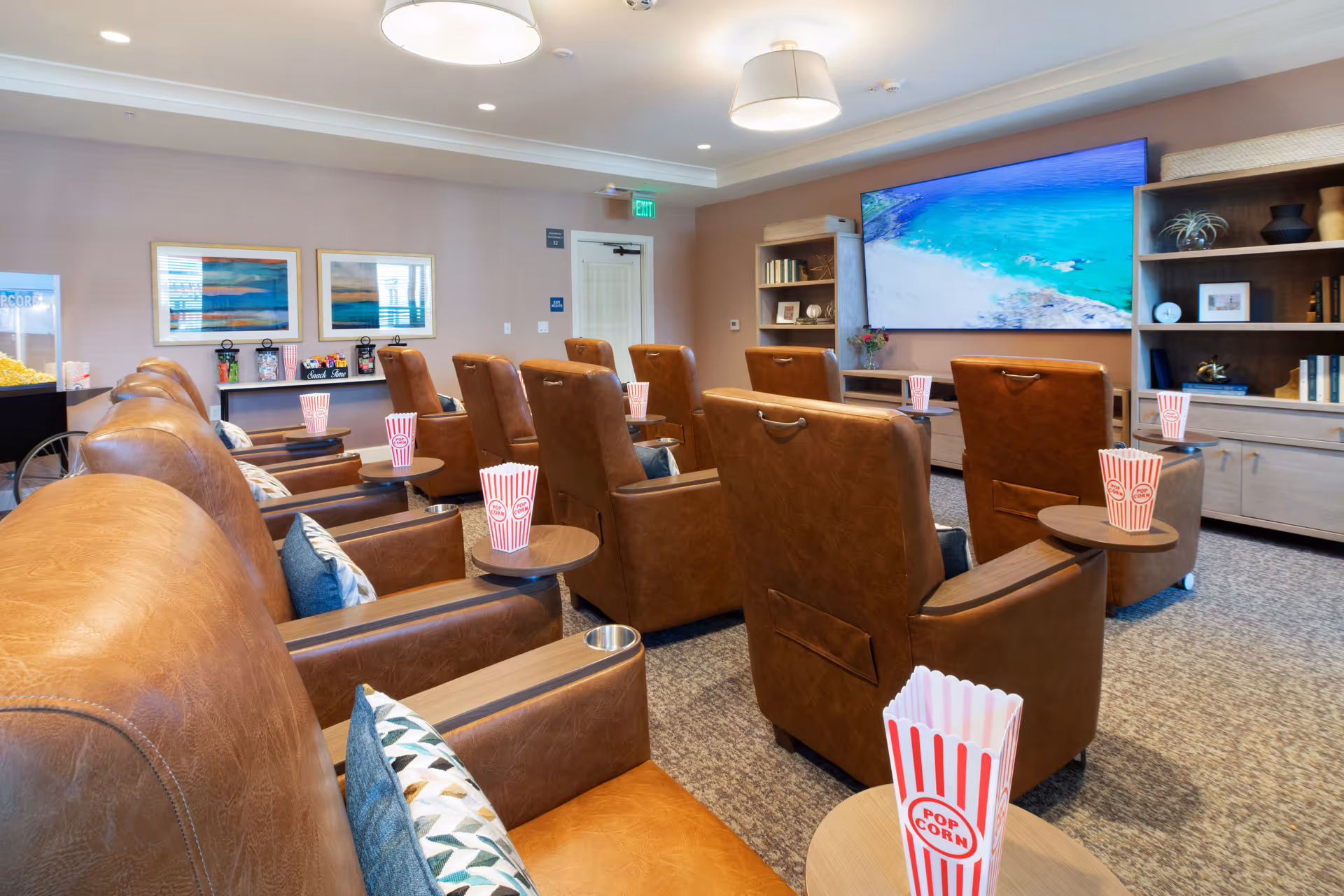 A cozy movie theater room with multiple brown leather recliner chairs arranged in rows, each with a small side table holding a popcorn container. A large flat-screen TV displays a beach scene. The room has soft lighting, carpeted floor, and shelves with books and decorative items.